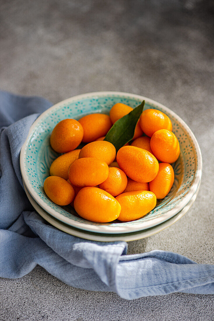 Schale mit frischen, leuchtenden Kumquats mit grünem Blatt von oben, auf einer strukturierten Oberfläche mit blauer Serviette