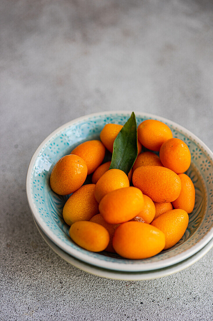 Draufsicht auf eine lebendige Schale frischer Kumquats mit grünem Blatt, platziert auf einer strukturierten grauen Oberfläche