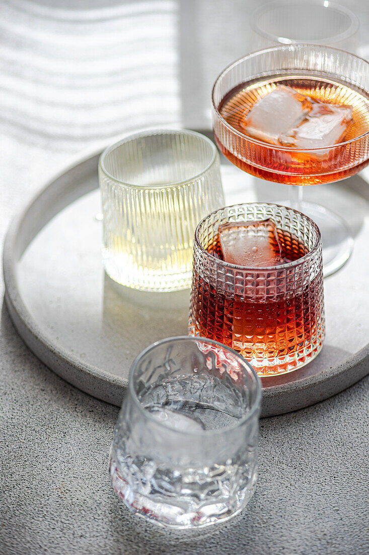 An assortment of alcoholic beverages on a round tray, each glass uniquely showcases a different drink, including cognac with cinnamon, limoncello with a sprig of rosemary, cherry liqueur over ice, and a clear, effervescent apple cider alongside a rich, iced plum liqueur