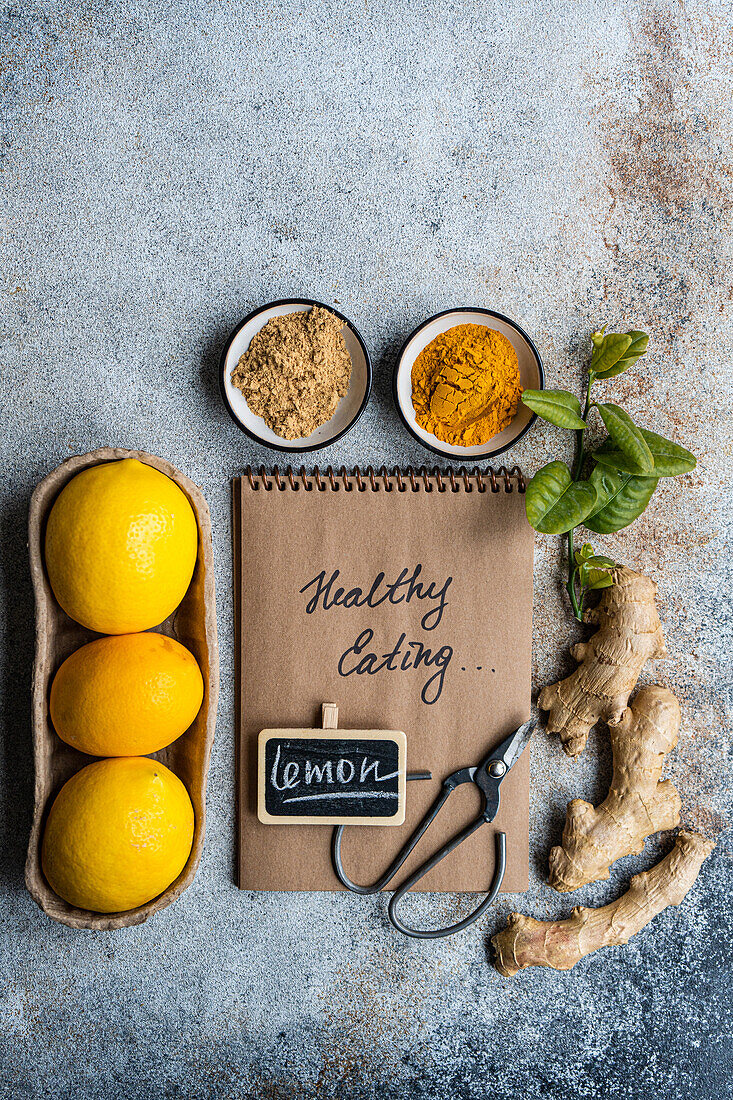 A notepad labeled healthy Cooking with a pen, fresh lemons, ginger, basil, and spices on a textured table