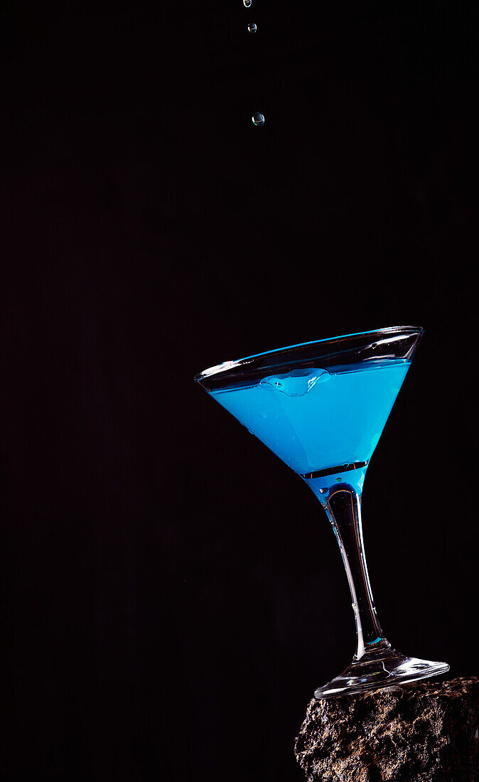 Drops of blue Lagoon cocktail pouring in crystal elegant glass placed on rough surface against black background