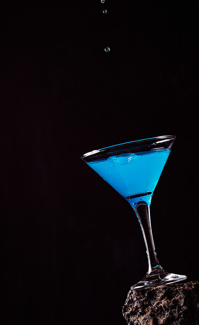 Tropfen eines blauen Lagunencocktails in einem eleganten Kristallglas auf einer rauen Oberfläche vor einem schwarzen Hintergrund