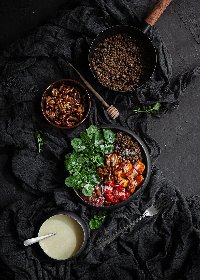 Draufsicht auf Eintopf mit Linsen neben Salat mit Kürbis und Paprika, dekoriert mit grünen Basilikumblättern auf schwarzem Hintergrund neben Salatdressing