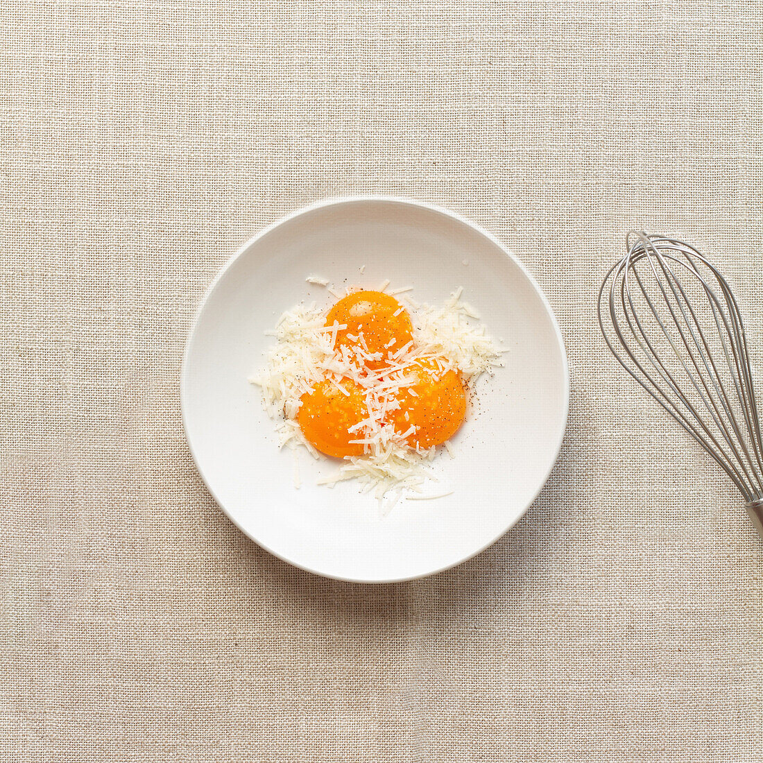From above two raw egg yolks nestled in grated cheese with a whisk on the side, ready for making Spaghetti Carbonara.