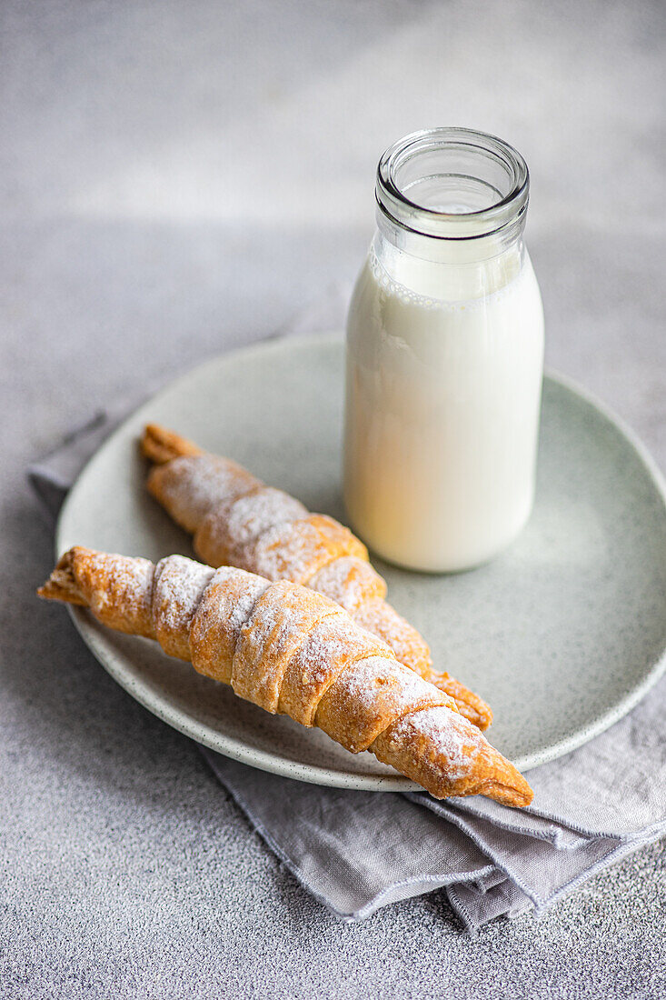 Zwei mit hausgemachter Marmelade gefüllte und mit Puderzucker bestäubte Gebäcktüten, begleitet von einer Flasche Milch, auf einem Keramikteller über einer grauen Serviette und Arbeitsplatte