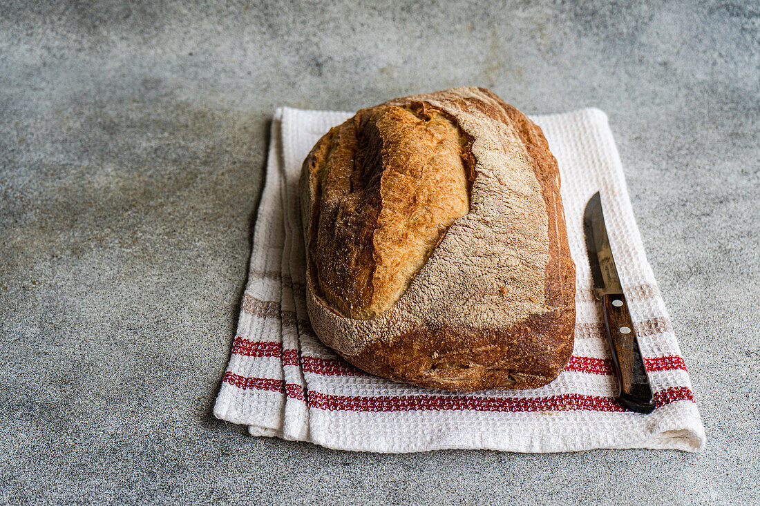 Draufsicht auf goldbraunes Roggensauerteigbrot auf einem weißen Tuch mit roten Streifen, daneben ein Brotmesser auf einer strukturierten Oberfläche