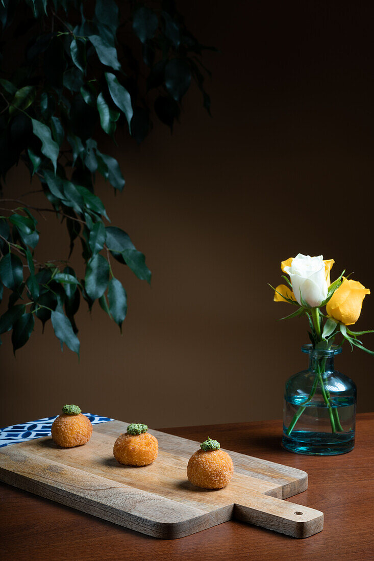 Drei goldene Arancini-Bällchen aus Reis und Käse, garniert mit Kräutern, präsentiert auf einem rustikalen Holzbrett, mit einer Vase mit gelben Rosen im Hintergrund.