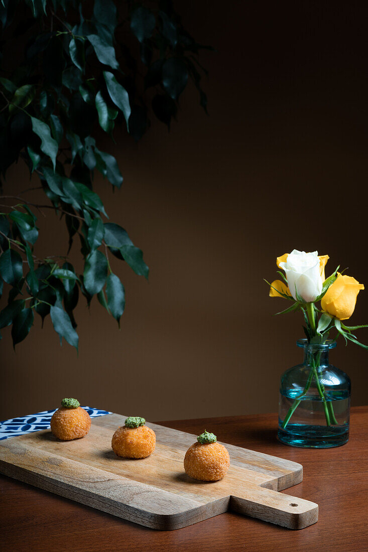Three golden rice and cheese arancini balls garnished with herbs, presented on a rustic wooden cutting board, with a vase of yellow roses in the backdrop.