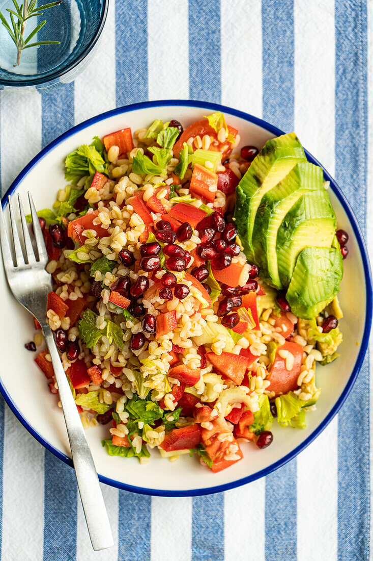 Draufsicht auf einen bunten Teller mit Bulgursalat, Avocado, Granatapfel und Gemüsewürfeln auf einem blau-weiß gestreiften Tuch