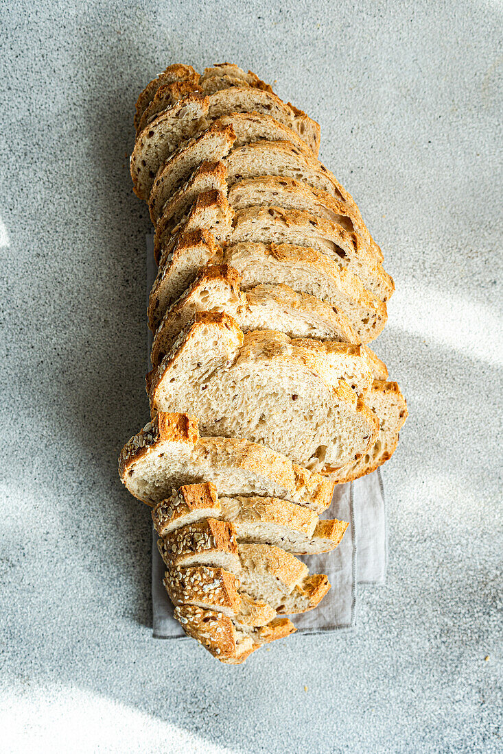 Top view of long loaf of seeded sourdough bread, sliced and arranged in a row on a linen cloth with a textured gray background