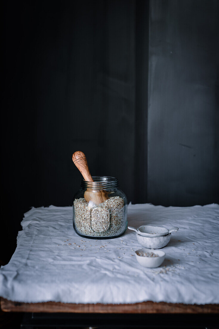 A rustic setup featuring homemade oatmeal stored in a glass jar with a wooden scoop, placed on a textured cloth over a wooden surface