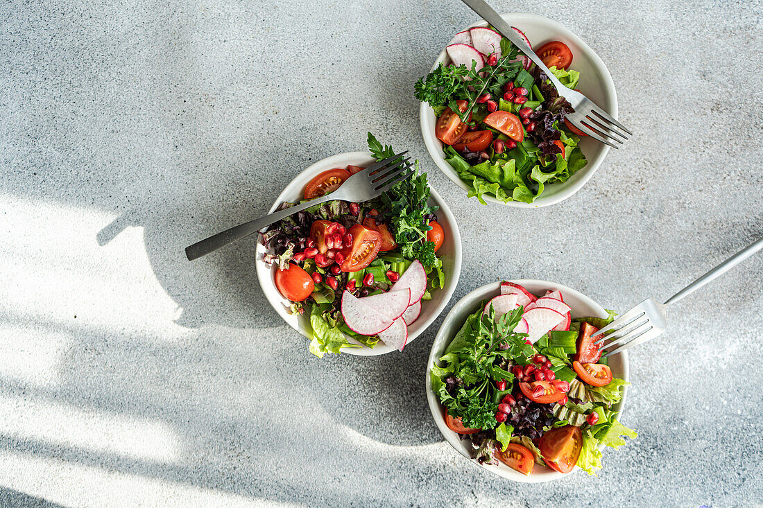 Draufsicht auf drei Schalen mit gesundem Gemüsesalat mit Kopfsalat, Rucola, Radieschen, Kirschtomaten und Granatapfelkernen vor einem Betonhintergrund