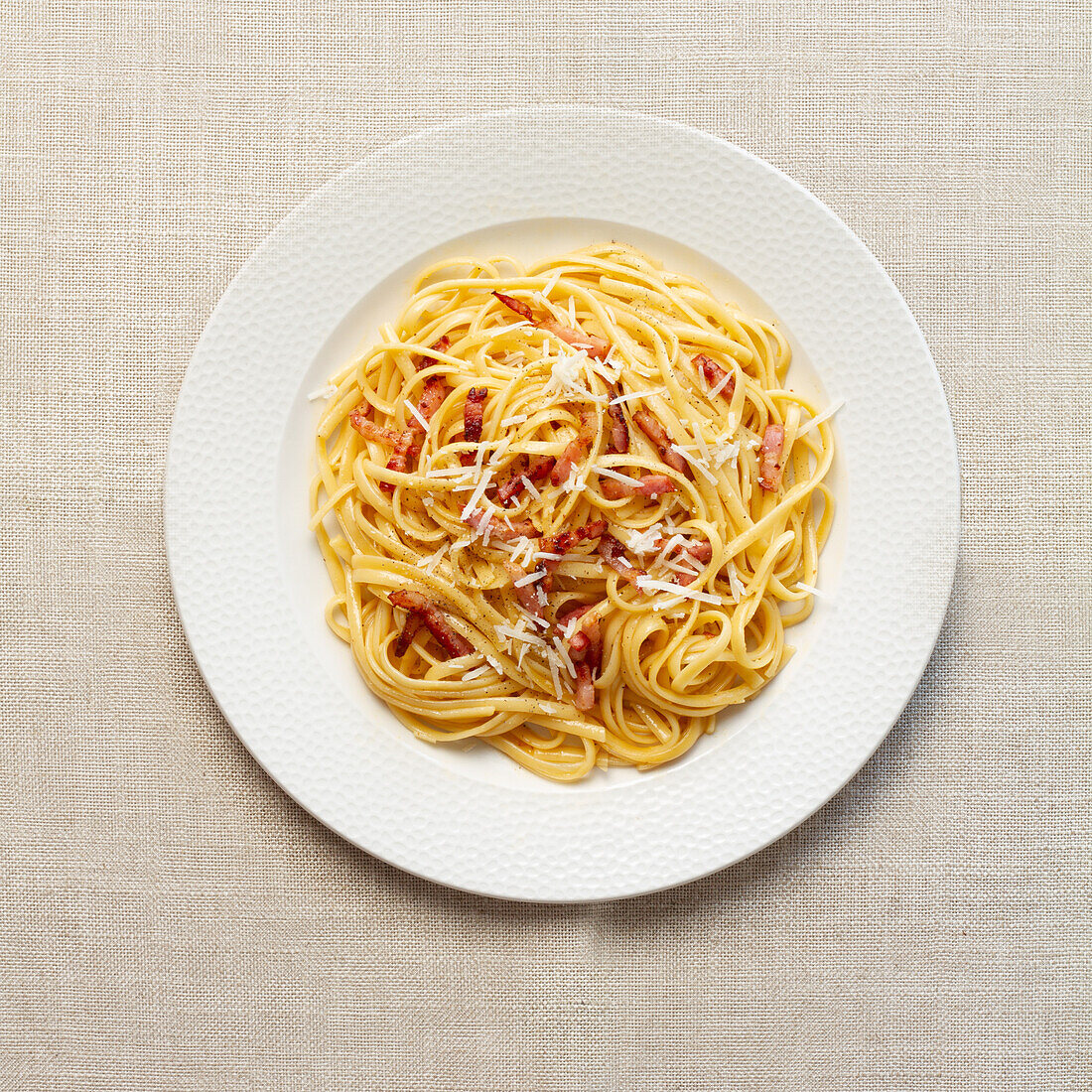 A top view of a classic Italian spaghetti carbonara, garnished with grated cheese and crispy bacon, served on a white textured background.