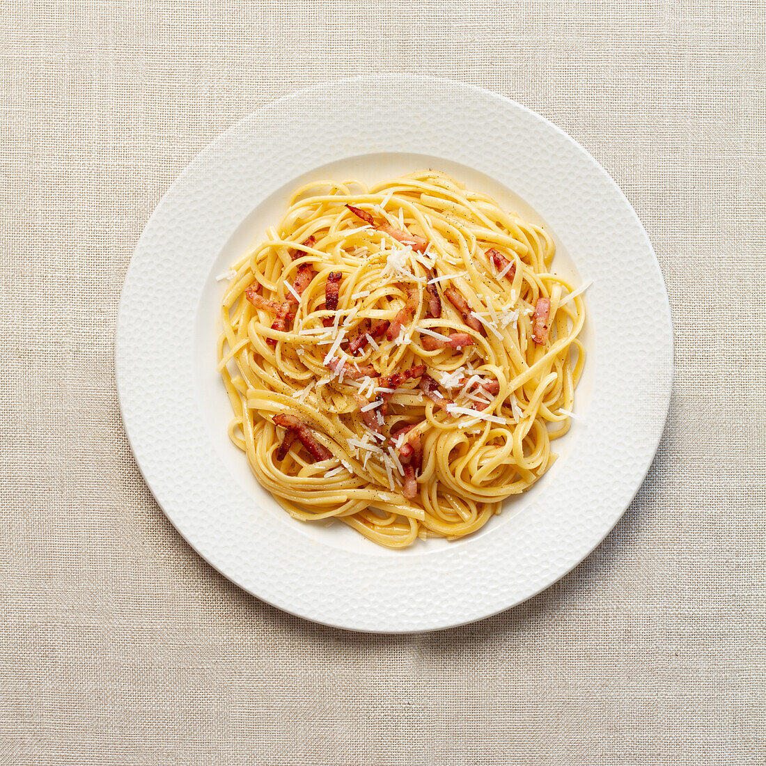 Eine Draufsicht auf klassische italienische Spaghetti Carbonara, garniert mit geriebenem Käse und knusprigem Speck, serviert auf einem weiß strukturierten Hintergrund.