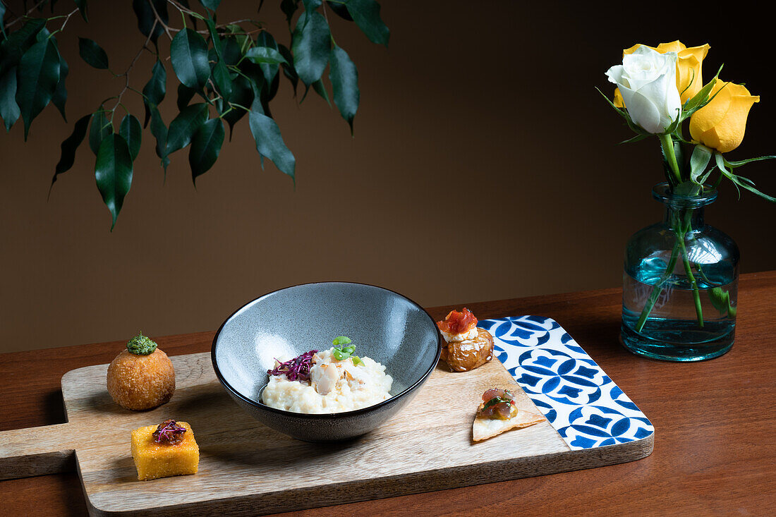 A variety of sophisticated canapés displayed on a wooden serving board, complemented by a bowl of creamy risotto and a vase of fresh yellow roses.