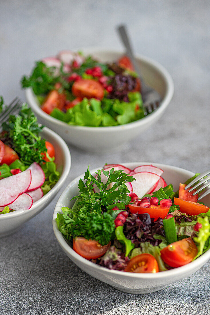 Freshly prepared vegetable salad featuring crisp lettuce, arugula, sliced radishes, cherry tomatoes, green onions, and pomegranate seeds in a white bowl on a textured grey surface