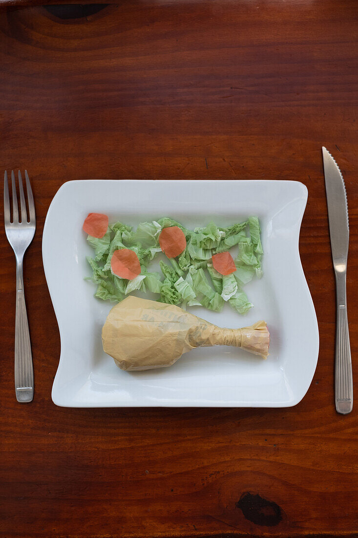 From above conceptual image of a plate with food items crafted from plastic bags, alongside real cutlery, on a wooden table.