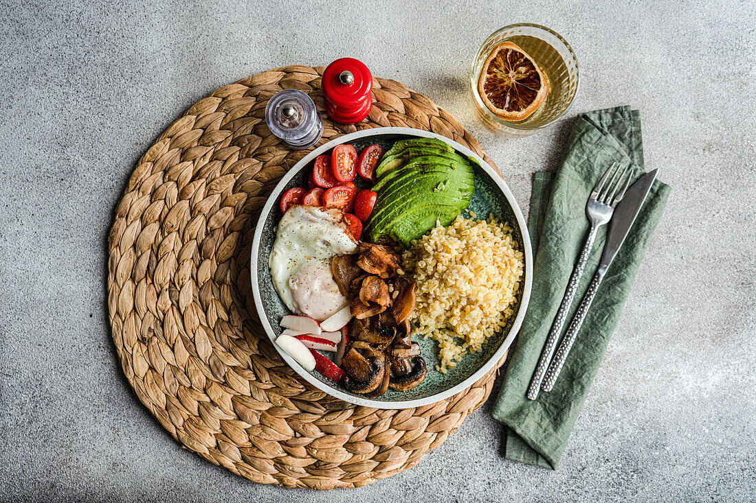 Draufsicht auf einen gesunden Teller mit Bulgur, Spiegelei, Avocado, Tomaten, Pilzen und Radieschen, mit Gewürzen und einem Glas Zitronenwasser vor einem strukturierten Hintergrund