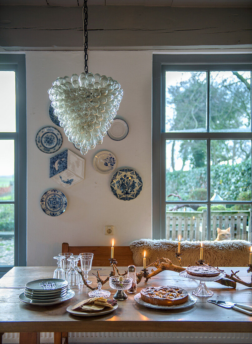 Set wooden table with candles and cake in front of large windows and wall decoration made of blue and white porcelain plates