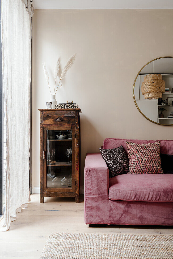 Pink sofa with patterned pillows next to rustic wooden cabinet and round wall mirror