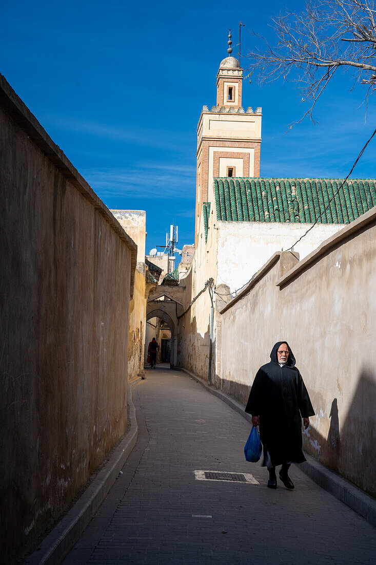 A man walks along a narrow street in Fez Medina with the minaret in the background on a clear day.