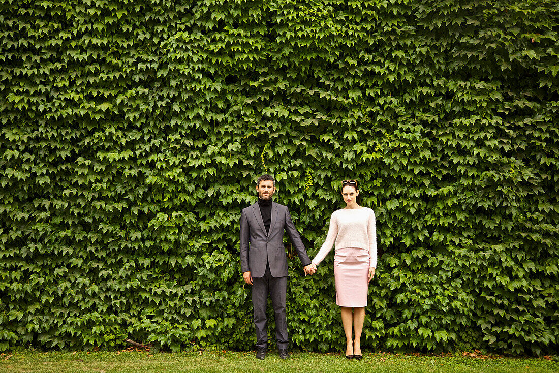 Händchenhaltendes Paar vor einer grünen Hecke