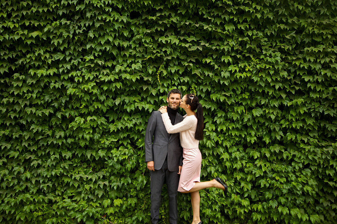 Woman Kissing Man on Cheek in front of Green Leafy Hedge