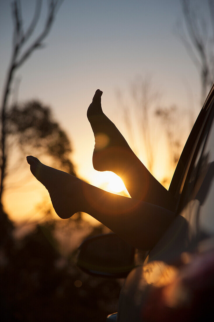 Füße einer Frau, die bei Sonnenuntergang aus dem Autofenster ragen