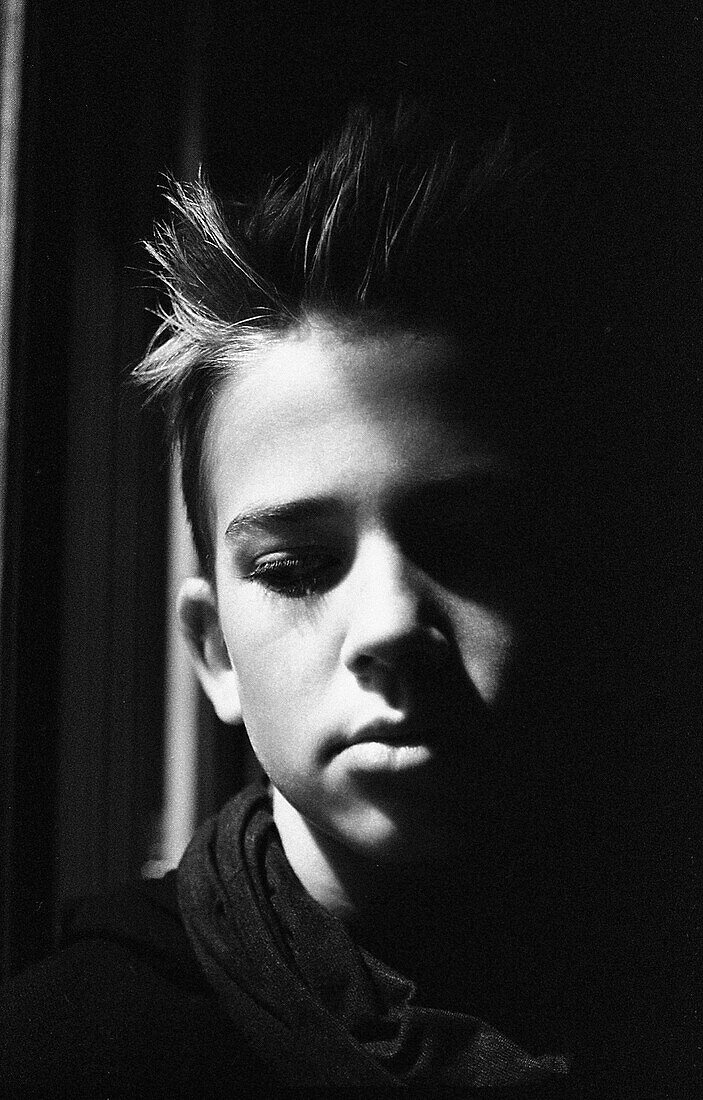 Head and shoulders portrait of contemplative teen boy looking down, face partially in shadow