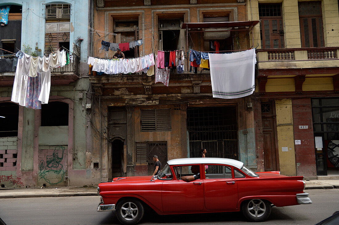 Kuba,Havanna,ein altes rotes amerikanisches Auto aus den 50er Jahren fährt an heruntergekommenen Häusern vorbei, in denen Wäsche trocknet