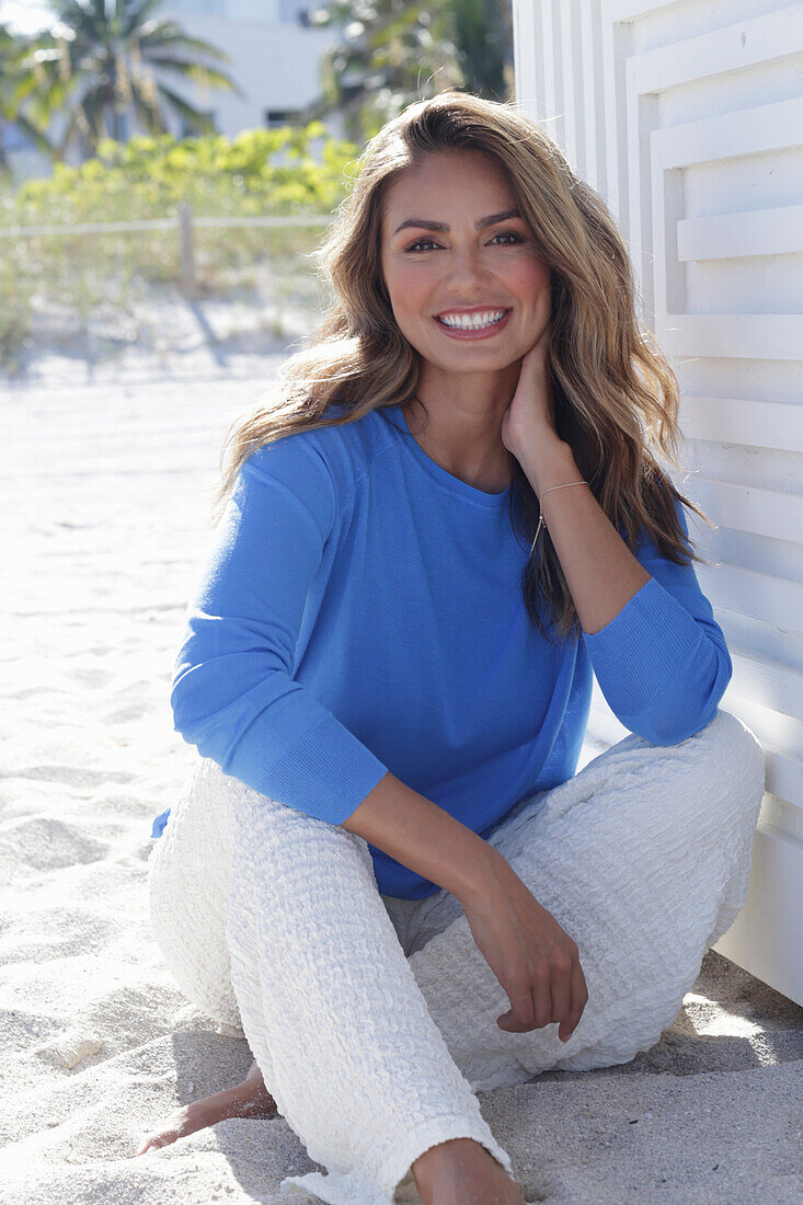 Frau in blauem Pullover und weißer Hose am Strand sitzend