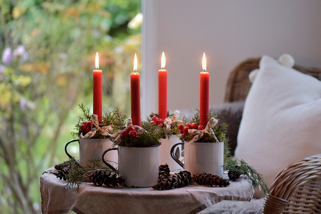 Four red Advent candles in enamel cups, decorated with fir branches and cones