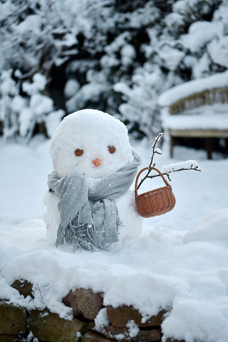 Schneemann mit grauem Schal und Korb im verschneiten Garten