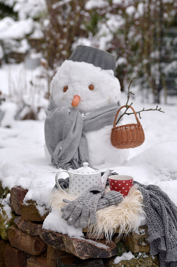 Snowman with grey scarf and basket, teapot and cups in the foreground