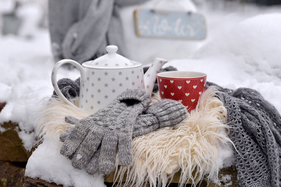 Teekanne und Tasse mit Handschuhen auf Felldecke im verschneiten Garten