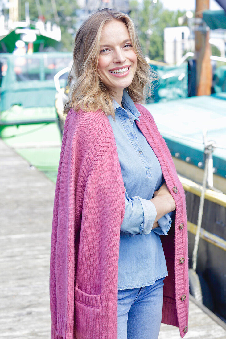 Junge Frau im Jeans-Outfit und rosa Strickjacke am Hafen