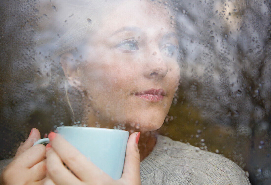 Frau hält Trinkbecher hinter verregnetem Fenster