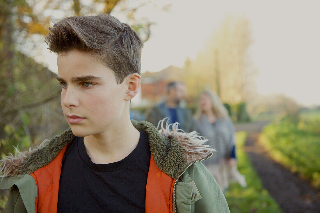 Teenage Boy Walking in Country Road, Parents in Background