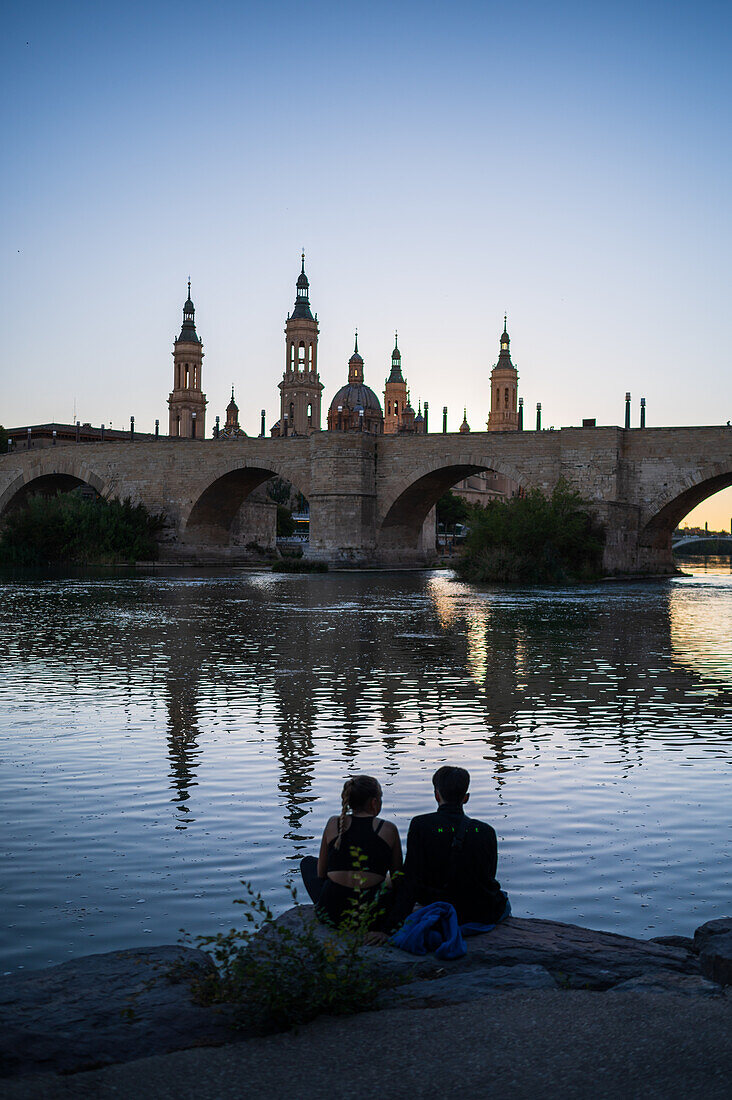 Junges Paar genießt den Sonnenuntergang am Ufer des Ebro, mit der Kathedrale-Basilika Unserer Lieben Frau von der Säule, Zaragoza, Spanien