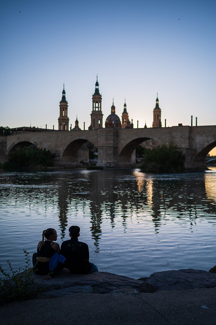 Junges Paar genießt den Sonnenuntergang am Ufer des Ebro, mit der Kathedrale-Basilika Unserer Lieben Frau von der Säule, Zaragoza, Spanien