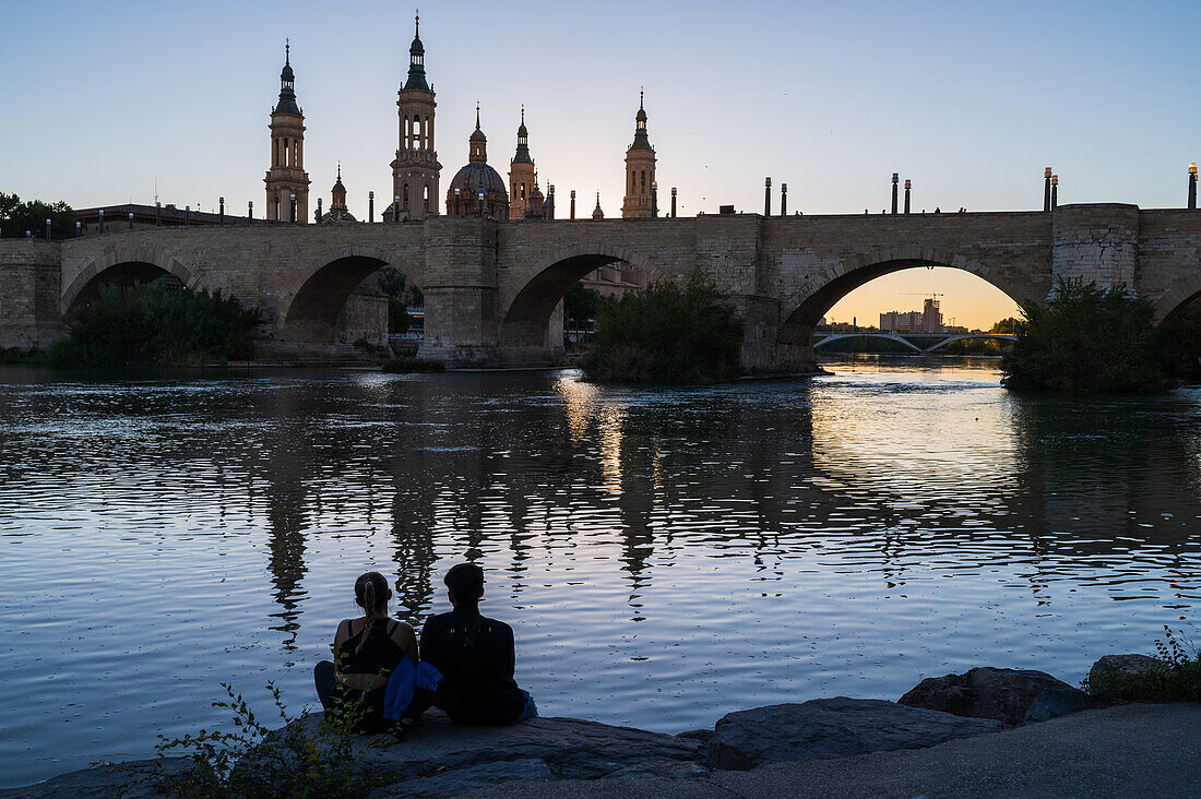 Junges Paar genießt den Sonnenuntergang am Ufer des Ebro, mit der Kathedrale-Basilika Unserer Lieben Frau von der Säule, Zaragoza, Spanien