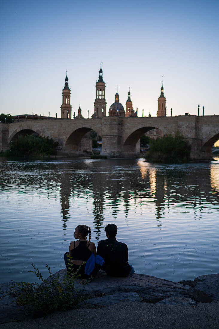 Young couple enjoying the sunset from the Ebro River bank, with Cathedral-Basilica of Our Lady of the Pillar, Zaragoza, Spain