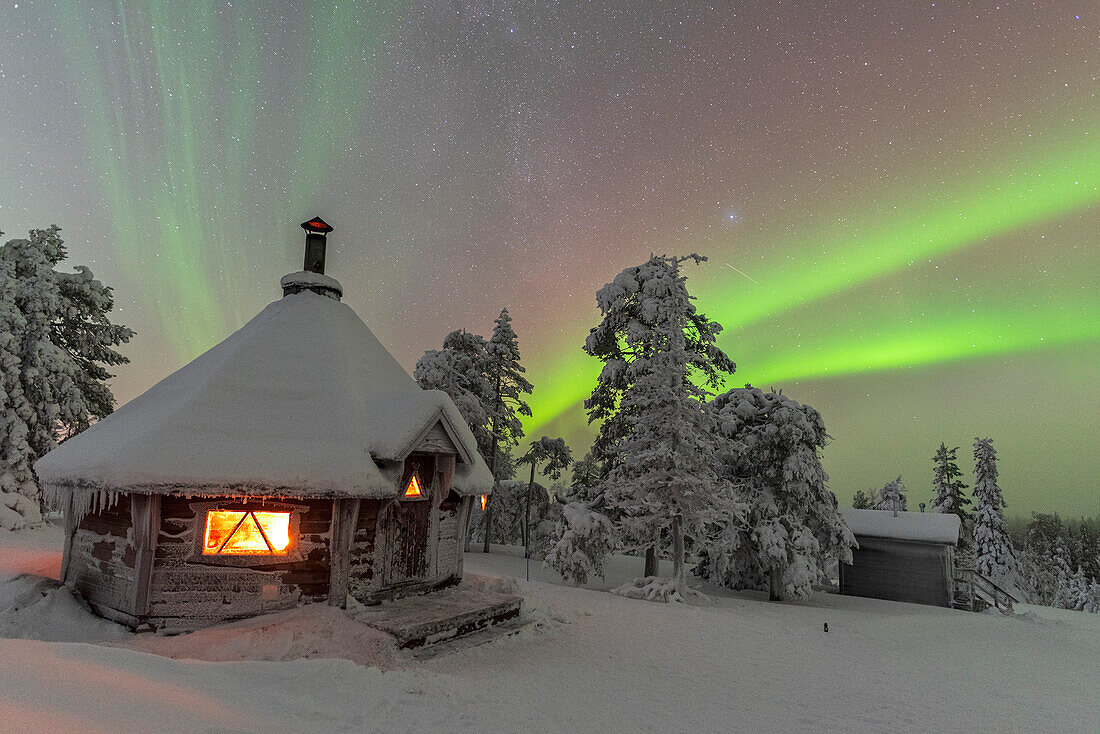 Grüntöne des Nordlichts (Aurora Borealis) über einer Schneelandschaft mit einer typischen, von einem Feuer beleuchteten Hütte im Vordergrund, Finnisch-Lappland, Finnland, Europa