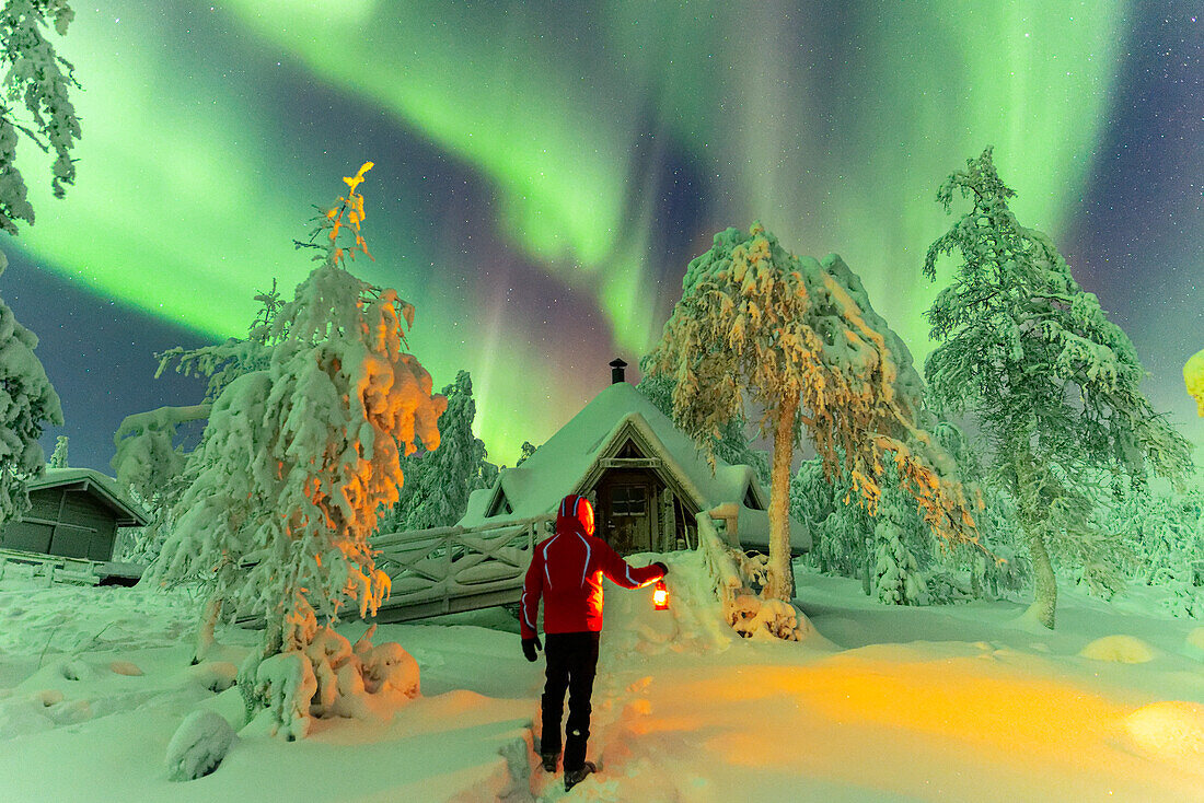 Mann mit Laterne vor einer typisch finnischen Hütte (kota) im verschneiten Wald unter Nordlicht (Aurora Borealis), Pallas-Yllastunturi-Nationalpark, Muonio, Finnisch-Lappland, Finnland, Skandinavien, Europa