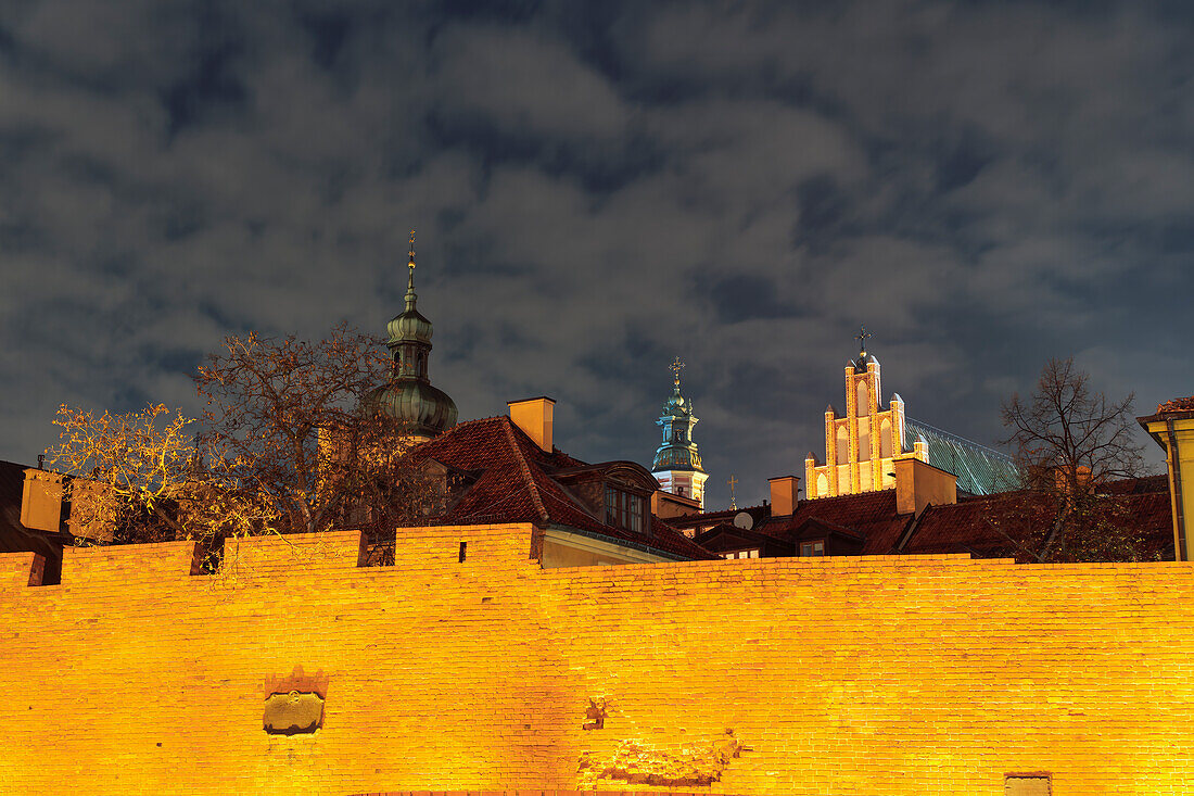 Night low angle view of illuminated defensive wall fortification in front of traditional low-rise roof-tiled houses and passing clouds above in the Old Town of Warsaw, Poland, Europe