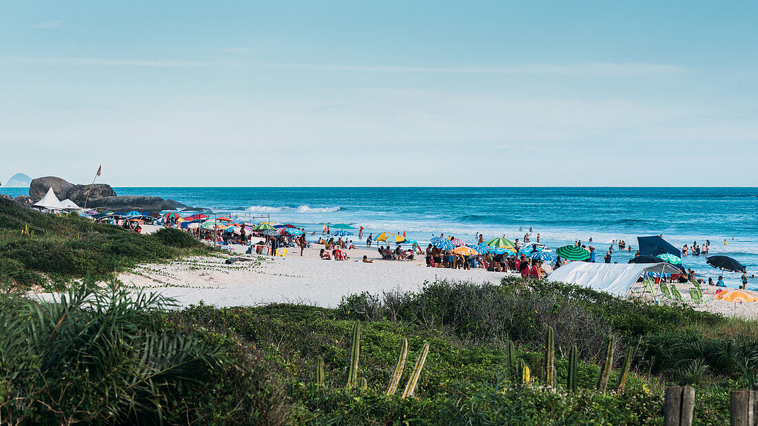 Crowds of beachgoers relax under colorful umbrellas on a sandy shoreline, taking in the ocean views and summer atmosphere, Grumari beach, Rio de Janeiro, Brazil, South America