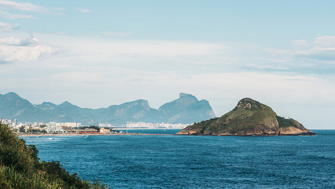 Malerischer Blick auf den Ozean mit üppiger grüner Insel und Stadtlandschaft vor Bergen an einem sonnigen Tag, mit Blick auf Pedra do Pombal und Barra da Tijuca, Rio de Janeiro, Brasilien, Südamerika