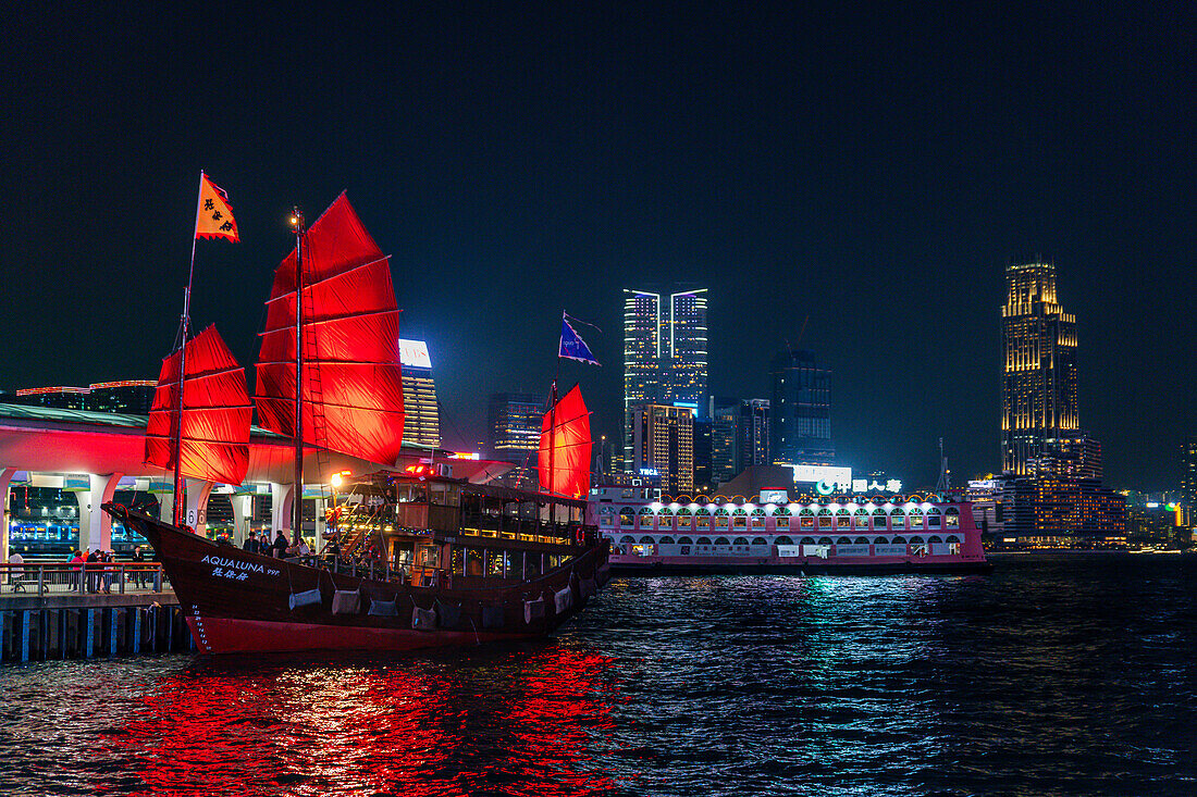 Traditionelles Segelboot mit roten Segeln bei Nacht, dahinter Hochhäuser im Zentrum von Hongkong, China, Asien