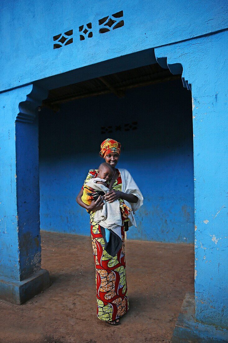 Ruanda, Landesmitte, Frau in buntem Lendenschurz mit ihrem Baby im Arm vor der Wand eines blauen Hauses