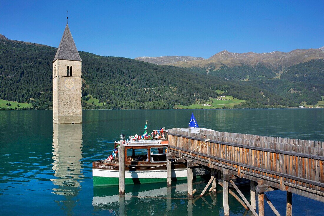 Italien, autonome Provinz Bozen, Reschensee, an einem Ponton vertäutes Boot mit verschlungenem Kirchturm, der aus dem Wasser des Sees auftaucht, umgeben von üppigen Bergen